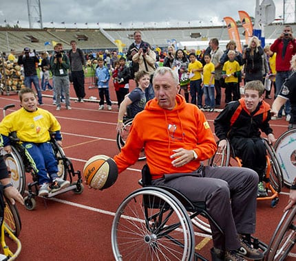 Johan Cruyff in een rolstoel met een basketbal in zijn handen en omringt door kinderen